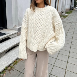 May include: Cream-colored cable knit sweater with a crew neck. The sweater features a diamond pattern on the front and cable knit detailing on the sleeves. The model is wearing beige pants. The sweater is a cozy, warm garment.