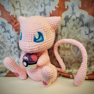 May include: A handmade, light pink crochet Mew plushie with blue eyes and a long tail. The plushie is holding a purple and white Pokeball. The crochet toy is a popular character from the Pokemon franchise.