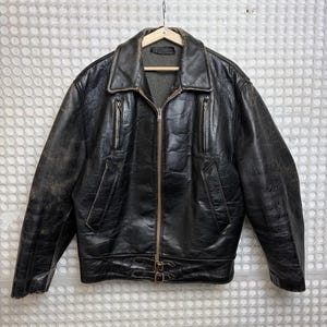 Puede incluir: Chaqueta de cuero negra con acabado envejecido, con cuello con solapas, cremallera frontal y múltiples bolsillos. La chaqueta tiene un detalle de cinturón en la cintura y está colgada de una percha de madera.