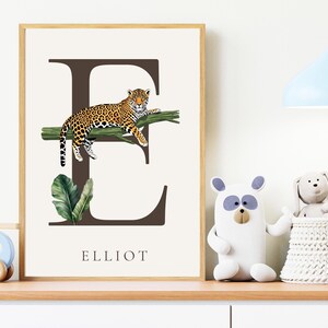Puede incluir: Impresión enmarcada con la letra "E" marrón y un leopardo sobre una rama verde. El nombre "ELLIOT" está debajo. Una hoja verde está en la parte inferior izquierda. El fondo es blanquecino. La impresión está en un marco de madera.