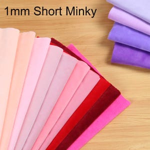 Può includere: Una collezione di campioni di tessuto minky corto da 1 mm disposti a ventaglio e in una pila separata. I colori includono varie tonalità di rosa, rosso e viola. Il testo 1mm Short Minky è stampato in nero nella parte superiore dell'immagine.