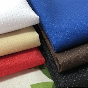 Può includere: Una pila di campioni di tessuto colorati in bianco, beige, rosso, blu, marrone e nero. Ogni tessuto ha una superficie strutturata con piccoli punti distanziati uniformemente. I tessuti sono piegati, mostrando la loro varietà.
