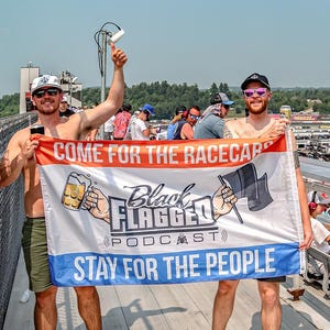 May include: Two men hold a banner with the text "COME FOR THE RACECARS, STAY FOR THE PEOPLE" and the "Black Flagged Podcast" logo. The banner has red, white, and blue stripes. The background shows a racetrack and spectators.