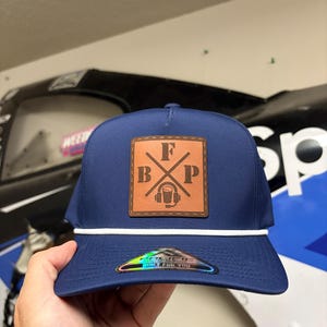 Könnte beinhalten: Eine marineblaue Baseballkappe mit einem weißen Kordeldetail an der Basis der Krone. Ein quadratischer, hellbrauner Lederaufnäher ist vorne mittig angebracht und zeigt die Buchstaben F, B und P sowie ein Headset-Symbol. Ein holografischer Aufkleber ist auf dem Schirm.