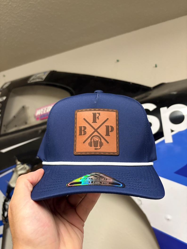 K&ouml;nnte beinhalten: Eine marineblaue Baseballkappe mit einem wei&szlig;en Kordeldetail an der Basis der Krone. Ein quadratischer, hellbrauner Lederaufn&auml;her ist vorne mittig angebracht und zeigt die Buchstaben F, B und P sowie ein Headset-Symbol. Ein holografischer Aufkleber ist auf dem Schirm.