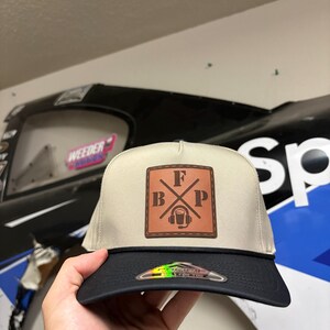 K&ouml;nnte beinhalten: Eine beige-schwarze Baseballkappe mit einem quadratischen braunen Lederaufn&auml;her auf der Vorderseite. Der Aufn&auml;her zeigt die Buchstaben F, B und P, die um eine zentrale Grafik eines Headsets mit Mikrofon angeordnet sind. Eine schwarze Kordel verl&auml;uft &uuml;ber den Schirm.