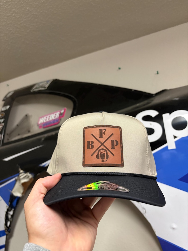 K&ouml;nnte beinhalten: Eine beige-schwarze Baseballkappe mit einem quadratischen braunen Lederaufn&auml;her auf der Vorderseite. Der Aufn&auml;her zeigt die Buchstaben F, B und P, die um eine zentrale Grafik eines Headsets mit Mikrofon angeordnet sind. Eine schwarze Kordel verl&auml;uft &uuml;ber den Schirm.