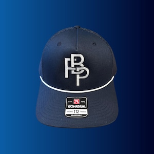 Puede incluir: Gorra de camionero azul marino con un logotipo blanco bordado y un borde blanco. La gorra tiene una parte trasera de malla y una correa ajustable. La etiqueta de la marca Richardson es visible.