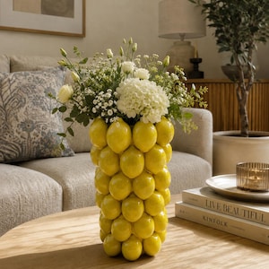 May include: A cylindrical ceramic vase featuring a textured exterior composed of numerous molded yellow lemons. The vase is filled with a bouquet of white flowers and green foliage, creating a bright, citrus-inspired decorative piece.