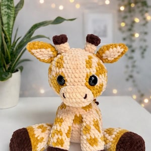 May include: A handmade crochet giraffe toy. The giraffe has a cream body with yellow and brown spots, large black eyes, and brown accents on the ears and hooves. The plush is sitting with its legs spread.