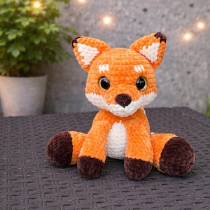 Könnte beinhalten: Ein handgefertigtes, orange-weißes Häkelfuchs-Plüschtier mit dunkelbraunen Akzenten. Der Fuchs hat große schwarze Augen und sitzt auf einer dunklen Oberfläche. Die Ohren haben weiße und braune Details. Ein süßes, kuscheliges Stofftier.