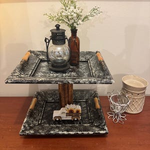 May include: A two-tiered decorative tray with a distressed black and white finish. It features a glass lantern, a brown bottle with white flowers, and a wooden truck with sunflowers. The tray has wooden handles.