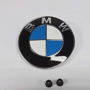May include: A BMW emblem in black, silver, and blue, featuring the letters "BMW". The circular design has a blue and white quadrant pattern. Two small black grommets are positioned below the emblem.