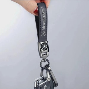 May include: A dark grey leather keychain strap featuring the Mercedes-Benz logo and text embossed in silver. The strap is attached to a metallic gunmetal-coloured hardware connector with a circular Mercedes-Benz emblem and a metal key ring.