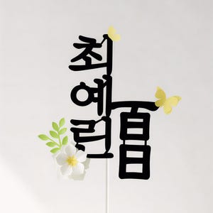 Peut inclure: Décoration de gâteau noire avec texte coréen, papillons et fleur. Le dessus est inséré dans un gâteau blanc. Le texte est en gras, police stylisée. Papillons jaunes et fleur blanche avec feuilles vertes.