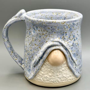 May include: A ceramic mug featuring a light blue glaze with dark blue and gold speckles. The mug has a sculpted design resembling a gnome with a smooth, light-colored nose protruding from beneath a draped hat brim and a textured white beard base.