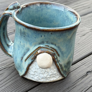 Puede incluir: Una taza de cerámica hecha a mano con un diseño de gnomo. La taza presenta un esmalte azul claro con detalles marrones y una barba blanca texturizada. El asa es curva y la taza tiene una forma única y caprichosa.