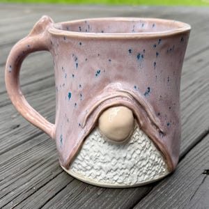 May include: A handmade ceramic mug with a gnome design. The mug features a pink glaze with blue speckles and a textured white beard. The handle is uniquely shaped, adding to its whimsical appeal.