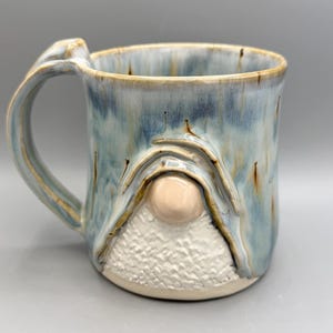 May include: A ceramic mug featuring a blue and tan glazed finish with a sculpted gnome design on the front. The gnome has a smooth, round, light-coloured nose and a textured, white, triangular beard area. It includes a curved handle on the left side.