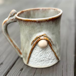 Peut inclure: Une tasse en céramique faite à la main avec un motif de gnome. La tasse présente un glaçage vert clair avec des accents bruns sur le bord et la poignée. Une sphère beige est centrée sur le devant, avec des lignes brunes formant un triangle.