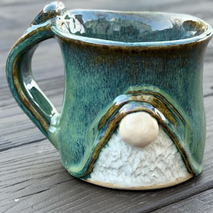 Puede incluir: Una taza de cerámica hecha a mano con un diseño de gnomo. La taza presenta un esmalte verde azulado con detalles marrones y una barba blanca texturizada. El asa es curva y la taza tiene una base redondeada. La taza mide aproximadamente 10 cm de alto.