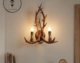 May include: A wall-mounted light fixture featuring a brown antler-shaped frame with two vertical candle-style bulbs. The fixture is centered on a plain wall, with a circular logo in the top left corner reading GrainGlazeStore Premium Chandeliers.