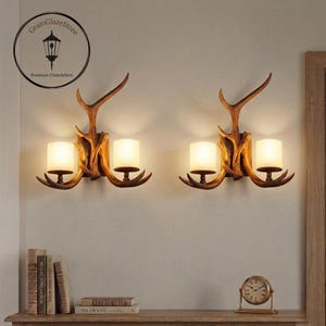 May include: Two antler-shaped wall sconces with warm, soft lighting. The sconces have a rustic, natural wood finish and are mounted on a light-colored wall. The image also shows a shelf with books and a small clock.