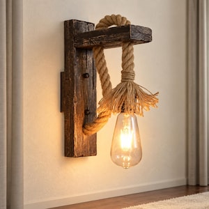 Pode incluir: Uma luminária de parede com uma base de madeira escura e envelhecida, apresentando uma corda grossa de juta em tom natural que desce do topo para segurar uma lâmpada transparente em forma de gota com um filamento brilhante visível.