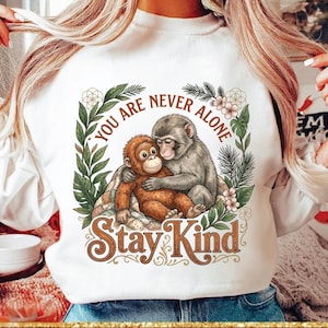 Pode incluir: Camisola branca com um gráfico de dois macacos abraçados, rodeados por flores e folhas. As palavras "YOU ARE NEVER ALONE" e "Stay Kind" estão escritas em uma fonte decorativa. Um estilo de roupa casual e confortável.