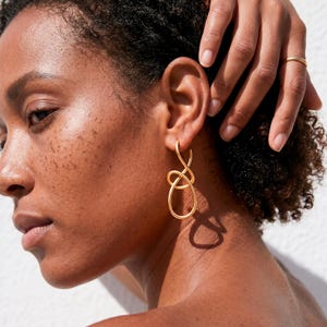 Könnte beinhalten: Goldfarbene, abstrakt geformte Ohrringe. Die Ohrringe haben ein geschlungenes Design mit glatter, polierter Oberfläche. Die Ohrringe werden am Ohr einer Person mit dunklen Haaren und Haut getragen. Ein goldener Ring ist am Finger der Person zu sehen.