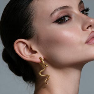 Puede incluir: Un pendiente dorado con forma de serpiente enroscada con una superficie texturizada similar a escamas. El diseño de la serpiente presenta un pequeño detalle de ojo oscuro y cuelga verticalmente del lóbulo de la oreja, mostrando un cuerpo fino.
