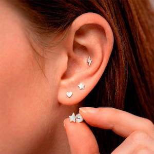 May include: A close-up of an ear with four small silver earrings: a lightning bolt, a star, a heart, and a star stud held by a hand. The earrings are simple and minimalist, suitable for everyday wear.