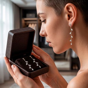 Puede incluir: Pendientes plateados con diseño de hojas, presentados en una caja de joyería marrón. Los pendientes presentan un diseño delicado y colgante con acentos claros y brillantes. La caja está abierta, revelando los pendientes sobre un interior de terciopelo oscuro.