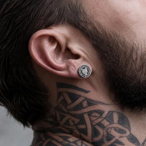 May include: A circular stud earring featuring a black and silver wolf head design surrounded by a border of runic-style symbols. The earring has a metallic finish and is worn in the earlobe, displaying a detailed engraved graphic motif.