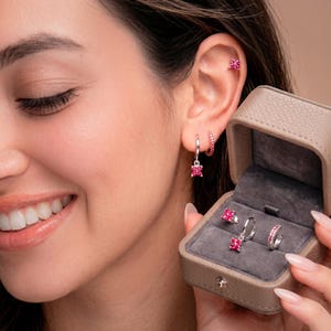 Puede incluir: Una mujer lleva un conjunto de pendientes de piedras preciosas rosas, que incluyen un estilo de botón, aro y colgante. Los pendientes se muestran en una pequeña caja de joyería gris abierta. La mujer sonríe y el fondo es de color neutro.