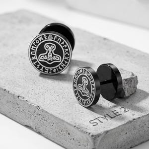 May include: A pair of black circular stud earrings featuring a white engraved Thor's hammer design surrounded by a border of runic symbols. The earrings have a flat front face and a post back. The text STYLE 2 is visible on the surface below.