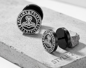 May include: A pair of black circular stud earrings featuring a white engraved Thor's hammer design surrounded by a border of runic symbols. The earrings have a flat front face and a post back. The text STYLE 2 is visible on the surface below.
