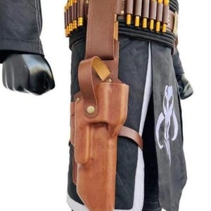 K&ouml;nnte beinhalten: Braune Lederholster mit passendem G&uuml;rtel, der eine Reihe von goldfarbenen Geschossen aufweist. Die Holster ist an einem schwarzen, gesteppten Kleidungsst&uuml;ck mit wei&szlig;em Besatz befestigt. Ein schwarzer Handschuh ist links zu sehen.