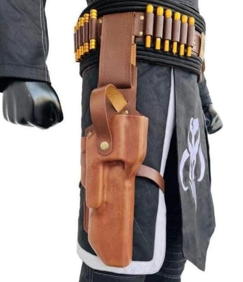 K&ouml;nnte beinhalten: Braune Lederholster mit passendem G&uuml;rtel, der eine Reihe von goldfarbenen Geschossen aufweist. Die Holster ist an einem schwarzen, gesteppten Kleidungsst&uuml;ck mit wei&szlig;em Besatz befestigt. Ein schwarzer Handschuh ist links zu sehen.