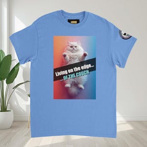 Könnte beinhalten: Hellblaues T-Shirt mit einem weißen Katzengrafik und dem Text "Living on the edge... OF THE COUCH." Die Katze befindet sich vor einem Farbverlauf aus Rot, Orange und Blau. Ein kleines Logo befindet sich auf dem Ärmel.