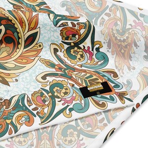 Könnte beinhalten: Nahaufnahme eines Stoffes mit einem farbenfrohen, verzierten Paisley-Design. Das Muster zeigt Wirbel, florale Elemente und stilisierte Blätter in Orange-, Türkis-, Rosa- und Goldtönen auf weißem Hintergrund. Ein schwarzes Etikett mit dem Wort "ENVERA" ist sichtbar.