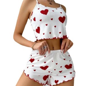 May include: White pajama set patterned with red hearts. The set comprises a camisole top with spaghetti straps and ruffled edges, and matching shorts. The top and shorts have red trim.