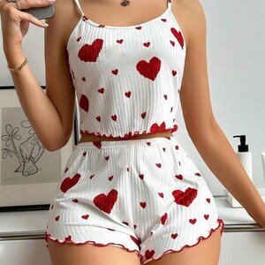 May include: White pajama set patterned with red hearts. The set comprises a camisole top with thin straps and matching shorts. Both the top and shorts feature red scalloped edging. The fabric has a ribbed texture.