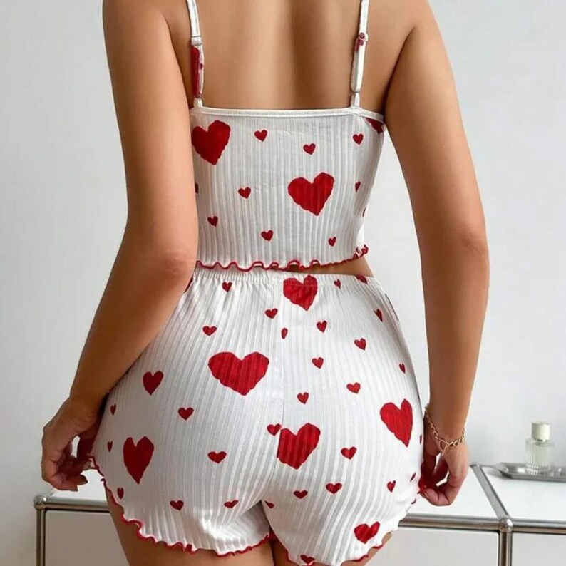 May include: White ribbed pajama set with a red heart pattern. The set includes a camisole top with adjustable straps and shorts. Both pieces have red scalloped edges. The fabric has a vertical ribbed texture.