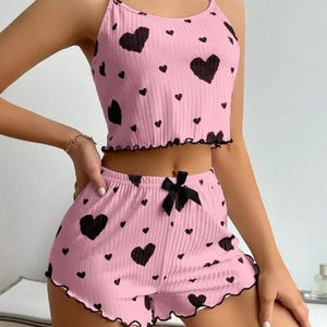May include: Pink pajama set with a black heart pattern. The set includes a camisole top and shorts with a black bow. The edges of the top and shorts have a black scalloped trim. The fabric has a ribbed texture.