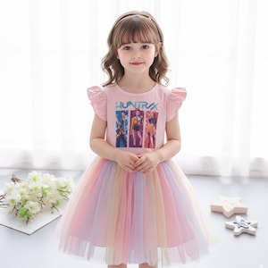 May include: A light pink dress with a rainbow tulle skirt. The top features the word "HUNTRAX" and a graphic of three figures. The dress has short, ruffled sleeves. The girl is wearing a headband.