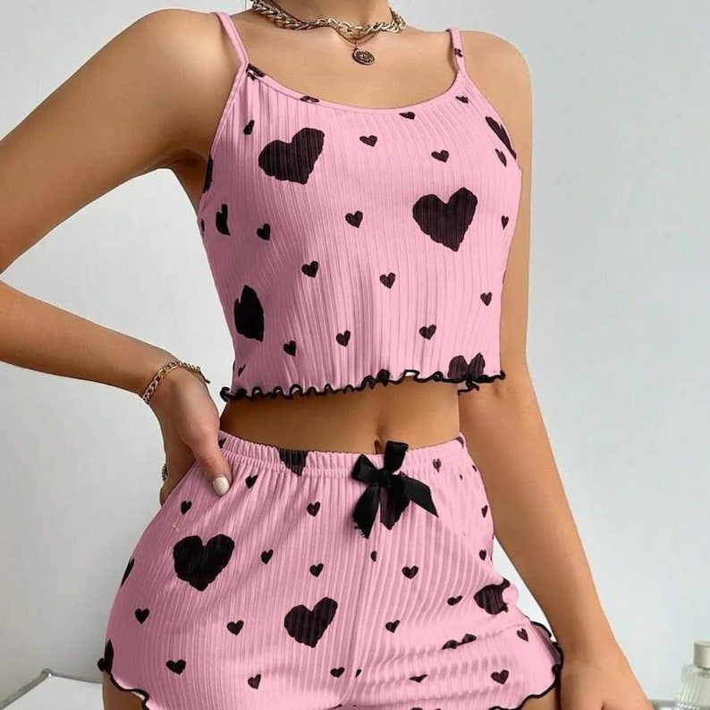 May include: A pink pajama set featuring a heart design. The set comprises a camisole top and shorts. The top has spaghetti straps and a frilled hem. The shorts have a black bow and a frilled hem. The hearts are black.