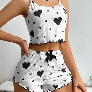 May include: White pajama set with black heart patterns. The set includes a camisole top with spaghetti straps and ruffled trim, and matching shorts with a black bow. The edges of the top and shorts are trimmed with black.