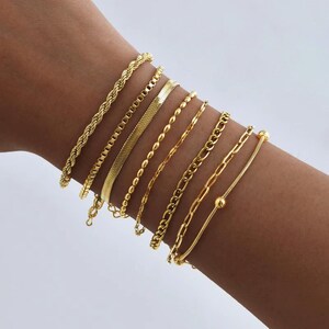 May include: An assortment of gold-coloured bracelets in diverse designs, including rope, chain, and beaded styles. The bracelets are displayed on a wrist, showcasing the different textures and patterns of the jewellery.