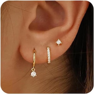 May include: An ear displaying three gold earrings. One is a small hoop with a dangling clear stone, another is a small hoop with clear stones, and the third is a small stud with a clear stone.